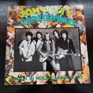 May include: A vintage album cover featuring Tom Petty and the Heartbreakers. The band is in a black and white photo, holding guitars. The album title is in teal and yellow, with the text "Rock 'N Roll Caravan '87" in green.
