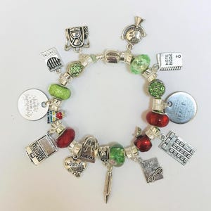 May include: A silver charm bracelet featuring various school-themed charms. Charms include a notebook, apple, calculator, pencil, and a heart that says "Favorite Teacher". The bracelet has green and red beads.