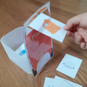 Puede incluir: Un cubo de plástico blanco con un gráfico rojo y naranja que dice "Plástico". Una mano sostiene una tarjeta con una ilustración de un contenedor naranja. Otras tarjetas con ilustraciones de varios objetos están esparcidas.