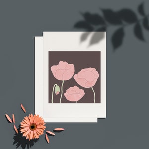 May include: A floral art print featuring three pink poppy flowers on a dark brown background. The print is on a white card, with a peach-colored gerbera daisy and petals in the corner. The image is set against a gray background.