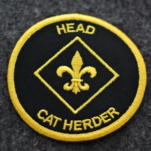 May include: A round, black patch with yellow embroidered text and accents. The text reads "HEAD" at the top and "CAT HERDER" at the bottom. A yellow fleur-de-lis is centered inside a yellow diamond.
