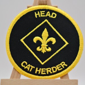 May include: Embroidered patch with a black circular background and a yellow border. The patch features the words "HEAD" and "CAT HERDER" in yellow, along with a yellow fleur-de-lis design inside a yellow diamond.