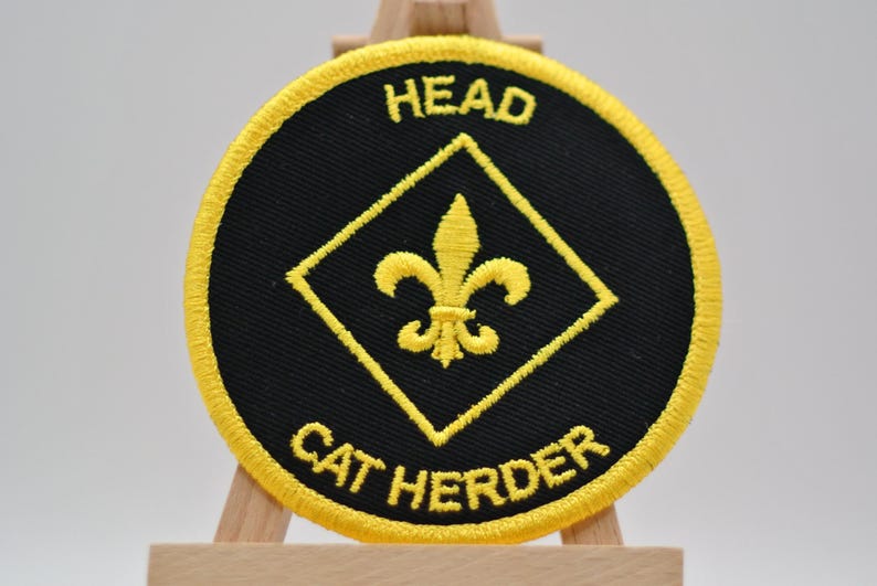 May include: Embroidered patch with a black circular background and a yellow border. The patch features the words "HEAD" and "CAT HERDER" in yellow, along with a yellow fleur-de-lis design inside a yellow diamond.