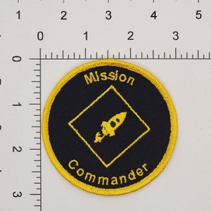 May include: A black and yellow round patch with the text "Mission Commander" and a yellow rocket ship inside a yellow diamond.