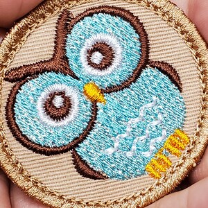 May include: Embroidered owl patch with a light blue body, brown accents, and white details. The owl has large, round eyes and is surrounded by a tan border. The patch is likely for clothing or accessories.