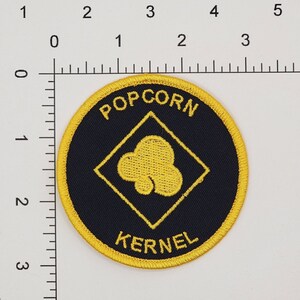 May include: A black and yellow embroidered patch with the words "POPCORN KERNEL" and a yellow popcorn kernel inside a yellow diamond.