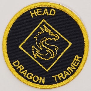 May include: Round black patch with gold embroidered text and design. The text reads "HEAD DRAGON TRAINER" around the edge. A gold dragon design is centered inside a gold diamond.