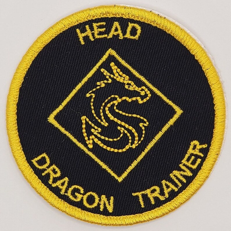 May include: Round black patch with gold embroidered text and design. The text reads "HEAD DRAGON TRAINER" around the edge. A gold dragon design is centered inside a gold diamond.