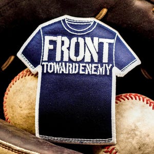 May include: A navy blue patch in the shape of a t-shirt with the words "FRONT TOWARD ENEMY" in white block letters. The patch has a white border and is set against a baseball glove and two baseballs.