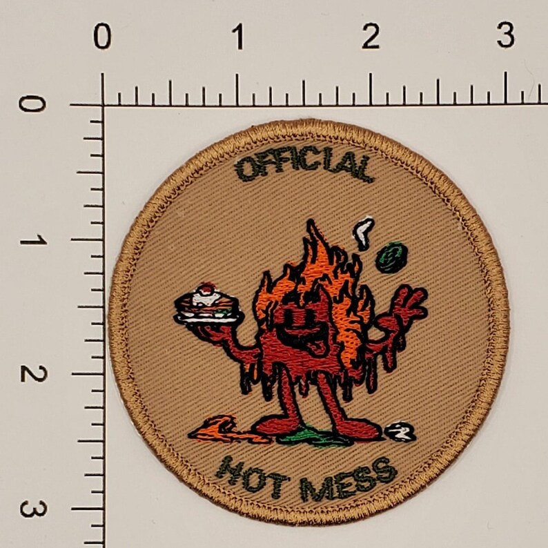 May include: A tan round patch with a cartoon character on fire holding a plate of food. The text "OFFICIAL HOT MESS" is on the patch.