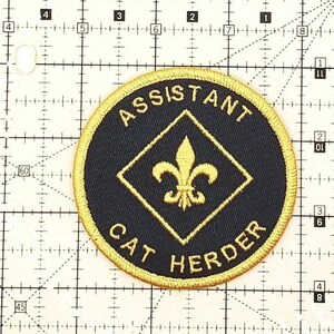 K&ouml;nnte beinhalten: Ein gestickter Aufn&auml;her in Schwarz und Gold mit dem Text "ASSISTANT CAT HERDER" und einer goldenen Fleur-de-lis in einer Raute.