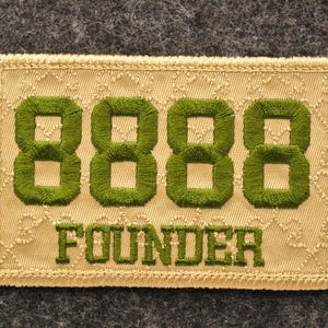 May include: Embroidered patch with the word "FOUNDER" below four green 8s. The patch is rectangular with a beige background and a decorative border. The background is a textured, patterned fabric.