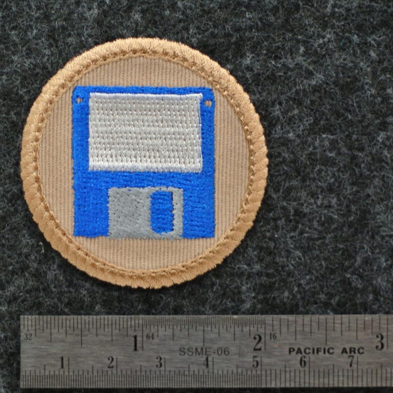 May include: A circular patch featuring a blue and silver floppy disk design on a tan background, surrounded by a tan border with a stitched edge. The patch is approximately 2 inches in diameter.