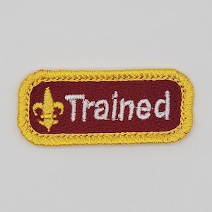 May include: A maroon rectangular patch with a yellow border and white stitching. The patch features a gold fleur-de-lis and the word "Trained" in white.