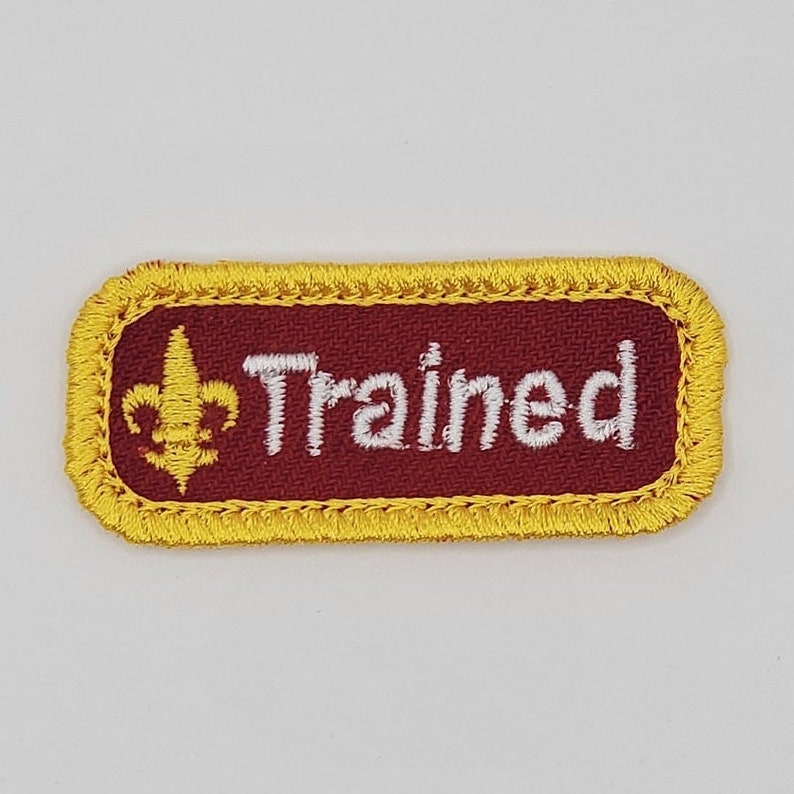 May include: A maroon rectangular patch with a yellow border and white stitching. The patch features a gold fleur-de-lis and the word "Trained" in white.