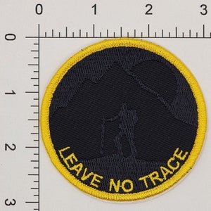 May include: Round black patch with a yellow border. The patch features a silhouette of a hiker with a walking stick against a mountain backdrop. The words "LEAVE NO TRACE" are in yellow.