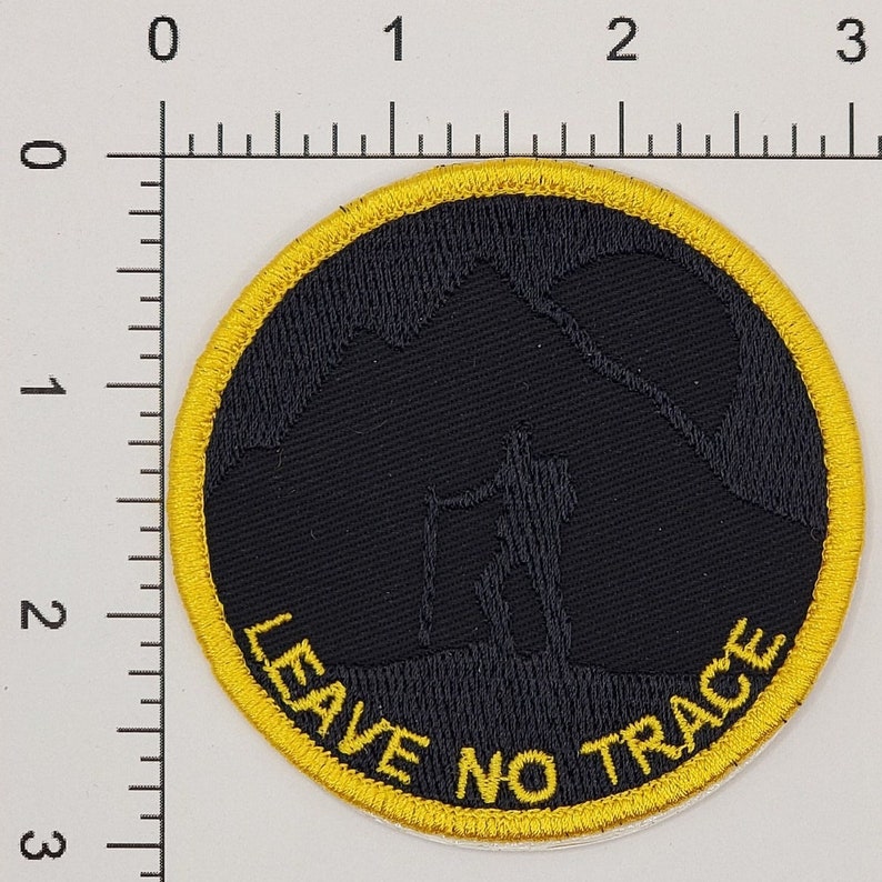 May include: Round black patch with a yellow border. The patch features a silhouette of a hiker with a walking stick against a mountain backdrop. The words "LEAVE NO TRACE" are in yellow.