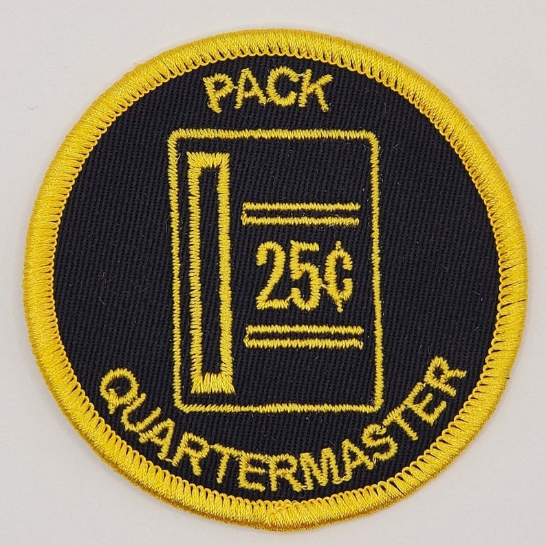 Pode incluir: Um remendo bordado amarelo e preto com o texto "PACK QUARTERMASTER" e uma imagem estilizada de uma m&aacute;quina de venda autom&aacute;tica com o texto "25&cent;".