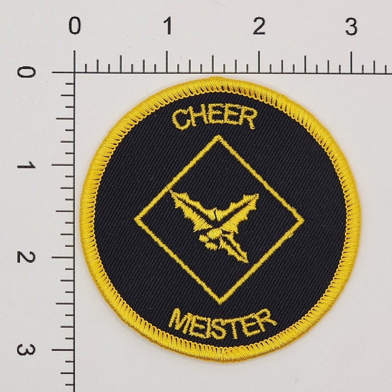 May include: A round, black and gold embroidered patch with the text "CHEER MEISTER" and a yellow holly leaf design inside a diamond shape.
