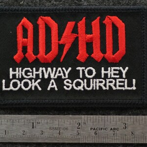 May include: Black embroidered patch with the text "AD/HD" in red gothic lettering, with a red lightning bolt. Below, white text reads "HIGHWAY TO HEY LOOK A SQUIRREL!". A ruler is at the bottom, showing inches.