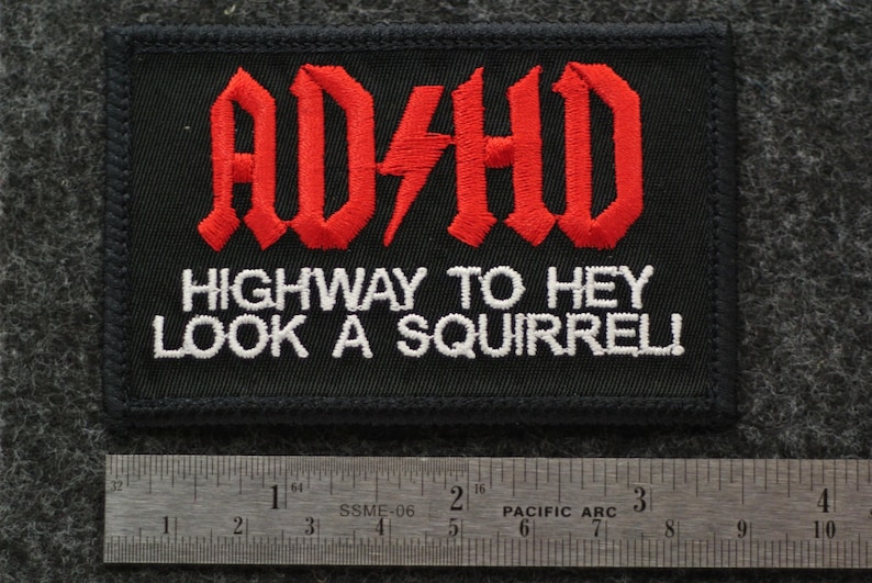 May include: Black embroidered patch with the text "AD/HD" in red gothic lettering, with a red lightning bolt. Below, white text reads "HIGHWAY TO HEY LOOK A SQUIRREL!". A ruler is at the bottom, showing inches.