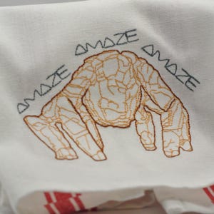 May include: White linen towel with an embroidered design. The design features a brown, abstract creature with the word "AMAZE" embroidered above and to the sides of the creature in gray thread. Red stripes are visible at the bottom.