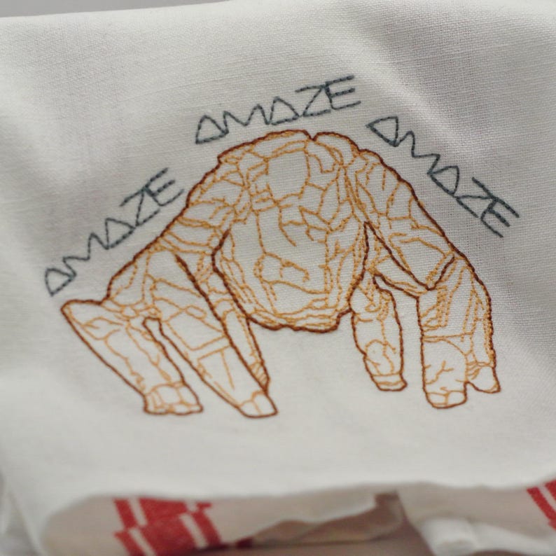 May include: White linen towel with an embroidered design. The design features a brown, abstract creature with the word "AMAZE" embroidered above and to the sides of the creature in gray thread. Red stripes are visible at the bottom.