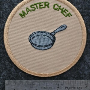 May include: A circular patch featuring the words "MASTER CHEF" in green above a blue frying pan. The patch has a beige background and a beige border with a stitched design.