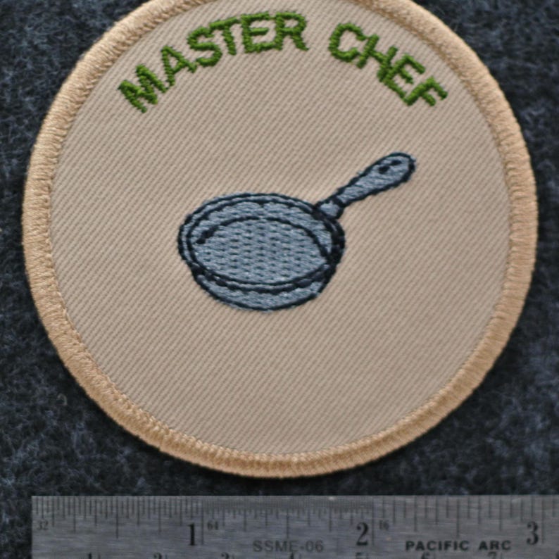 May include: A circular patch featuring the words "MASTER CHEF" in green above a blue frying pan. The patch has a beige background and a beige border with a stitched design.
