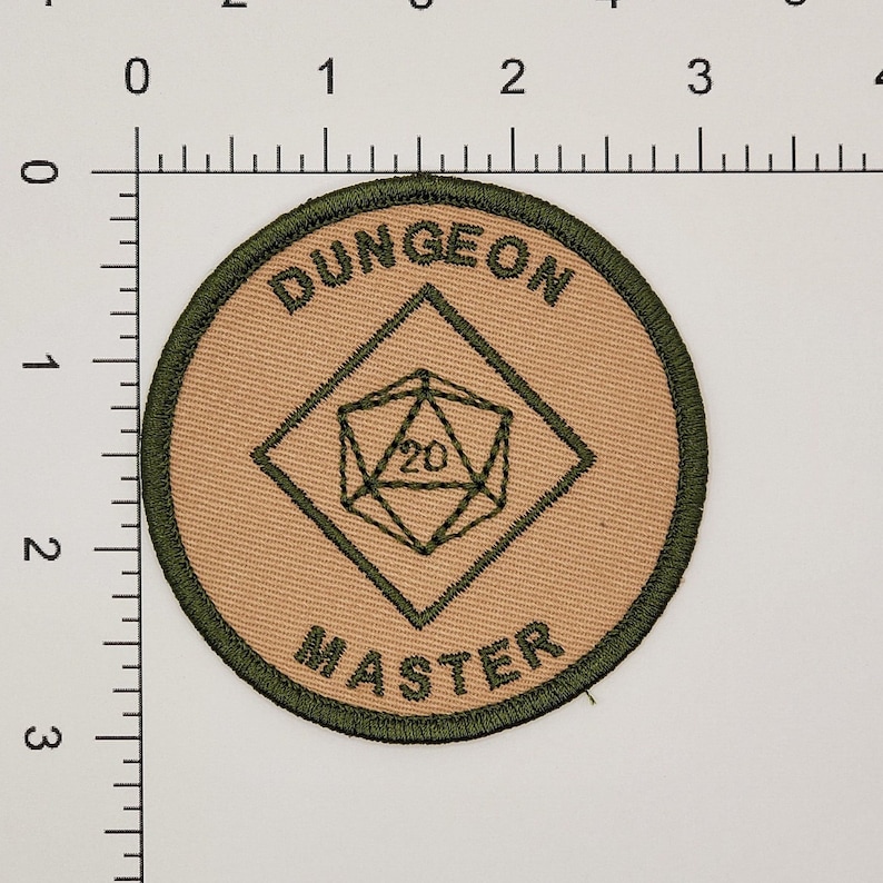 K&ouml;nnte beinhalten: Ein gestickter Aufn&auml;her in Tan und Gr&uuml;n mit dem Text "Dungeon Master" und einem 20-seitigen W&uuml;rfel in einer Diamantenform.