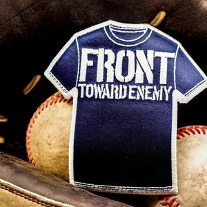 May include: A navy blue patch in the shape of a t-shirt with the words "FRONT TOWARD ENEMY" in white. The patch is set against a baseball glove and two baseballs.