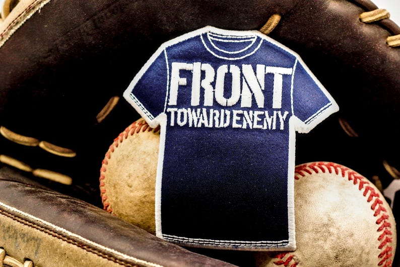 May include: A navy blue patch in the shape of a t-shirt with the words "FRONT TOWARD ENEMY" in white. The patch is set against a baseball glove and two baseballs.