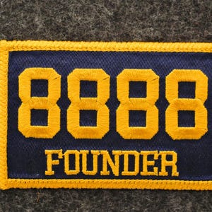 May include: A rectangular patch with a navy blue background and gold embroidered text and border. The number "8888" is prominently displayed above the word "FOUNDER". The patch is likely for a club or organization.