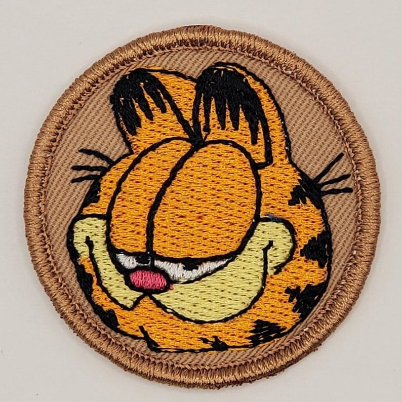 May include: Embroidered patch featuring Garfield the cat's face. The patch has a tan background with a brown border. Garfield's face is orange with black markings, a pink nose, and a yellow mouth.