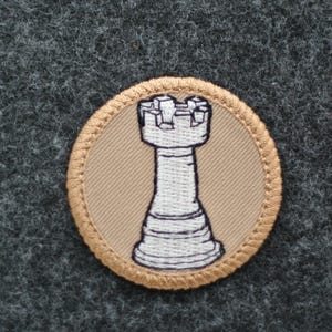 Op de afbeelding: Ronde geborduurde patch met een wit schaaktorenontwerp op een beige achtergrond. De patch heeft een beige rand met een gestikte rand, tegen een grijze, getextureerde stof.