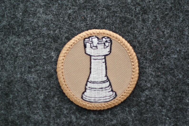 Op de afbeelding: Ronde geborduurde patch met een wit schaaktorenontwerp op een beige achtergrond. De patch heeft een beige rand met een gestikte rand, tegen een grijze, getextureerde stof.