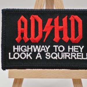 May include: Black embroidered patch with the text "ADHD" in red gothic lettering, a red lightning bolt, and the phrase "HIGHWAY TO HEY LOOK A SQUIRREL!" in white. The patch is displayed on a small wooden easel.