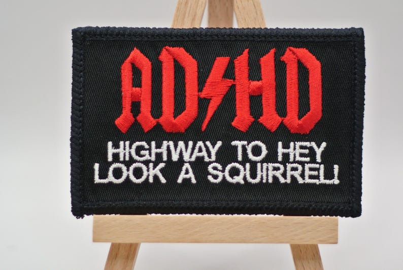 May include: Black embroidered patch with the text "ADHD" in red gothic lettering, a red lightning bolt, and the phrase "HIGHWAY TO HEY LOOK A SQUIRREL!" in white. The patch is displayed on a small wooden easel.