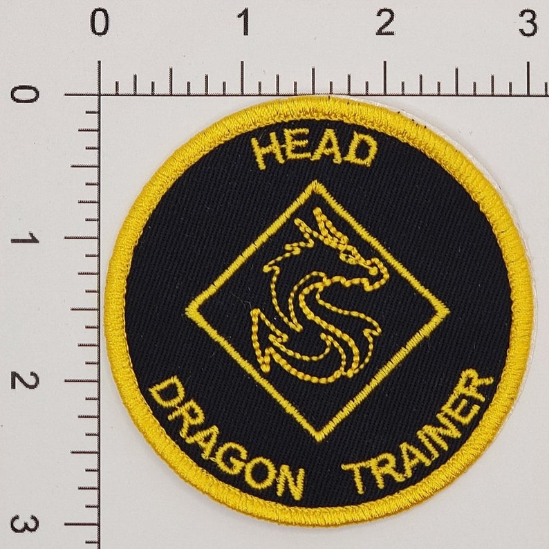 May include: Round black patch with yellow embroidered border and text. The patch features the words "HEAD DRAGON TRAINER" in yellow. A yellow dragon design is in the center, inside a yellow diamond.