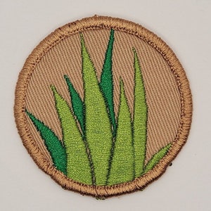 May include: A circular embroidered patch featuring a light brown background with a detailed green plant design. The plant has multiple long, pointed leaves. The patch is bordered with a brown stitched edge.