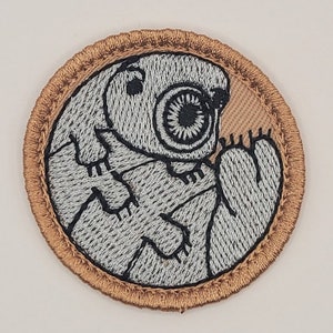 May include: Embroidered patch featuring a stylized creature with a large eye and clawed feet. The design is primarily light gray with black detailing, set against a tan background, and framed by a brown border.