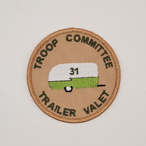 May include: A tan round patch with green embroidered text that reads "Troop Committee Trailer Valet". The center of the patch features a green and white camper with the number "31" on the side.