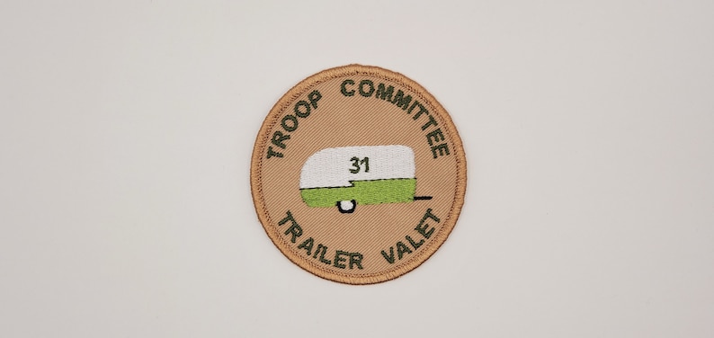 May include: A tan round patch with green embroidered text that reads "Troop Committee Trailer Valet". The center of the patch features a green and white camper with the number "31" on the side.