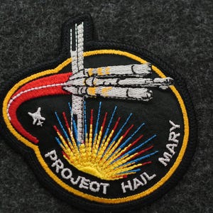 Puede incluir: Parche bordado con un dise&ntilde;o de nave espacial y el texto "PROJECT HAIL MARY". El parche tiene un fondo negro con una combinaci&oacute;n de colores amarillo, rojo, blanco y azul. Una estrella es visible en el lado izquierdo del parche.