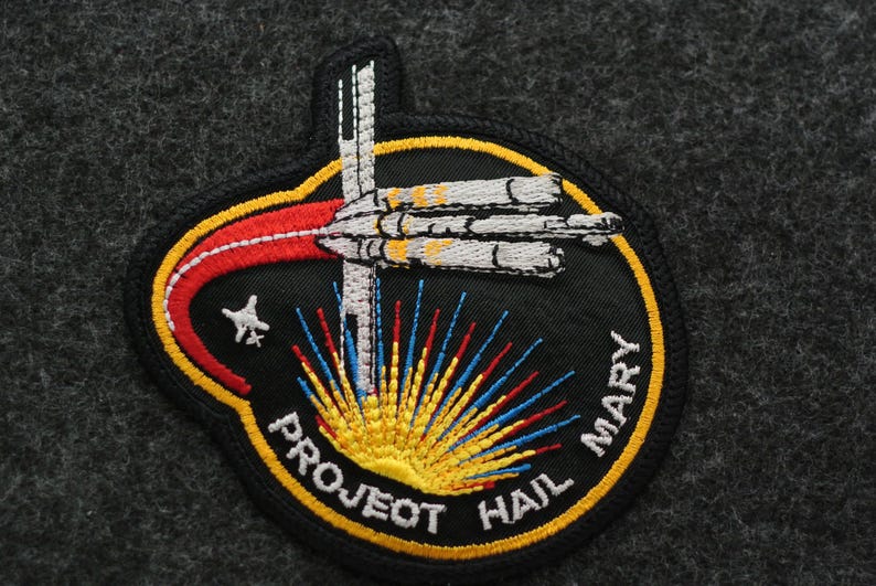 Puede incluir: Parche bordado con un dise&ntilde;o de nave espacial y el texto "PROJECT HAIL MARY". El parche tiene un fondo negro con una combinaci&oacute;n de colores amarillo, rojo, blanco y azul. Una estrella es visible en el lado izquierdo del parche.