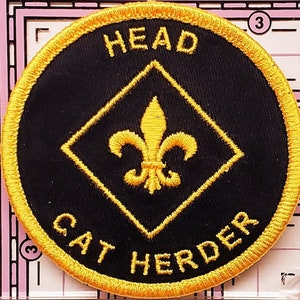 May include: Round black embroidered patch with gold trim and lettering. The text reads "HEAD" at the top and "CAT HERDER" at the bottom. A gold fleur-de-lis is centered inside a gold diamond.