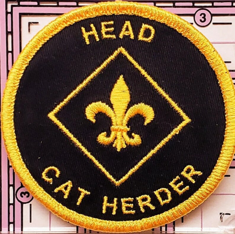 May include: Round black embroidered patch with gold trim and lettering. The text reads "HEAD" at the top and "CAT HERDER" at the bottom. A gold fleur-de-lis is centered inside a gold diamond.