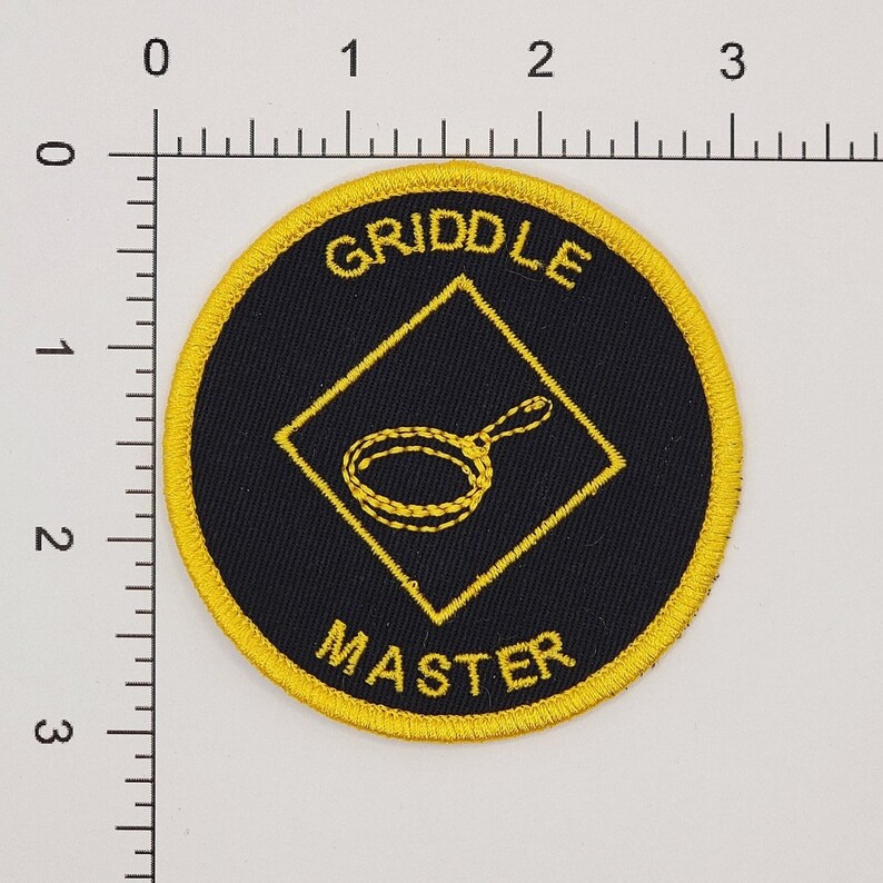 May include: A yellow and black embroidered patch with the text "Griddle Master" and a yellow diamond shape with a black frying pan inside.