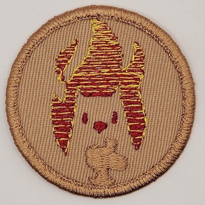 May include: Embroidered patch with a circular shape. The patch features a tan background with a detailed design of a stylized face and flame-like elements in red and yellow. The border is a stitched gold color.