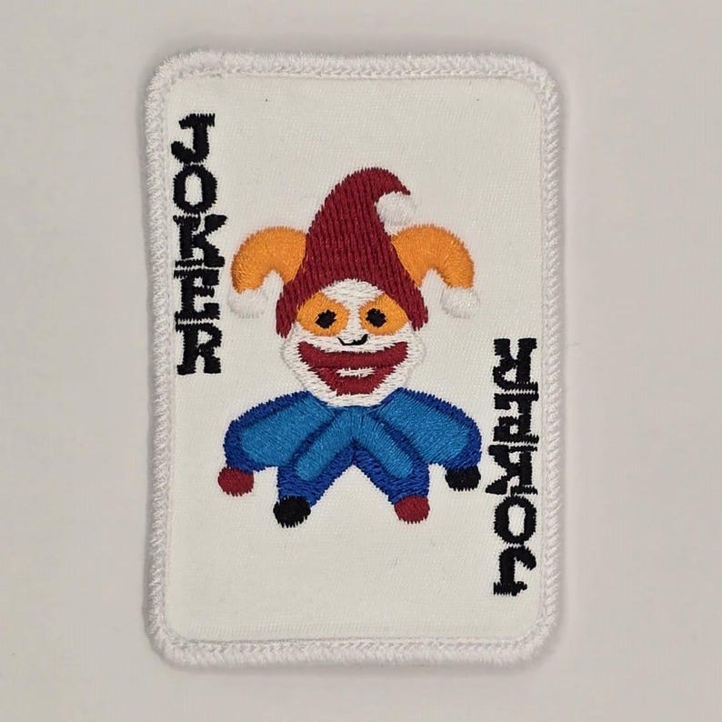 Op de afbeelding: Gestikt patch van een joker speelkaart met een witte achtergrond en zwarte tekst die "JOKER" zegt. De joker is afgebeeld als een cartoonfiguur met een rode hoed, oranje haar en een blauw-wit kostuum.